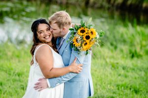 Bruidspaar omhelst elkaar in het gras tijdens hun trouwdag, met een zonnebloemboeket in de hand.